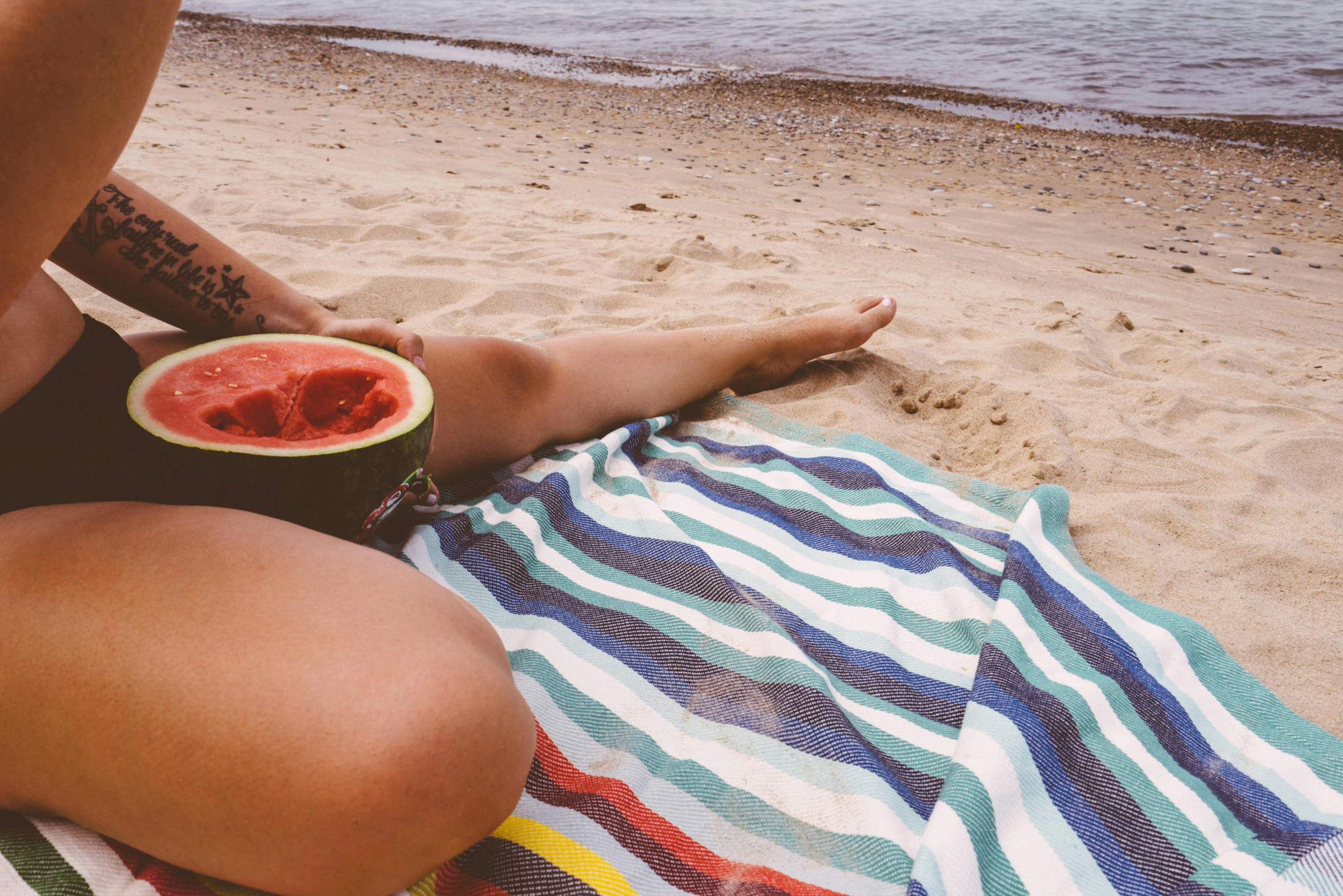 Person sitting on a beach with a watermelon on a striped towel.