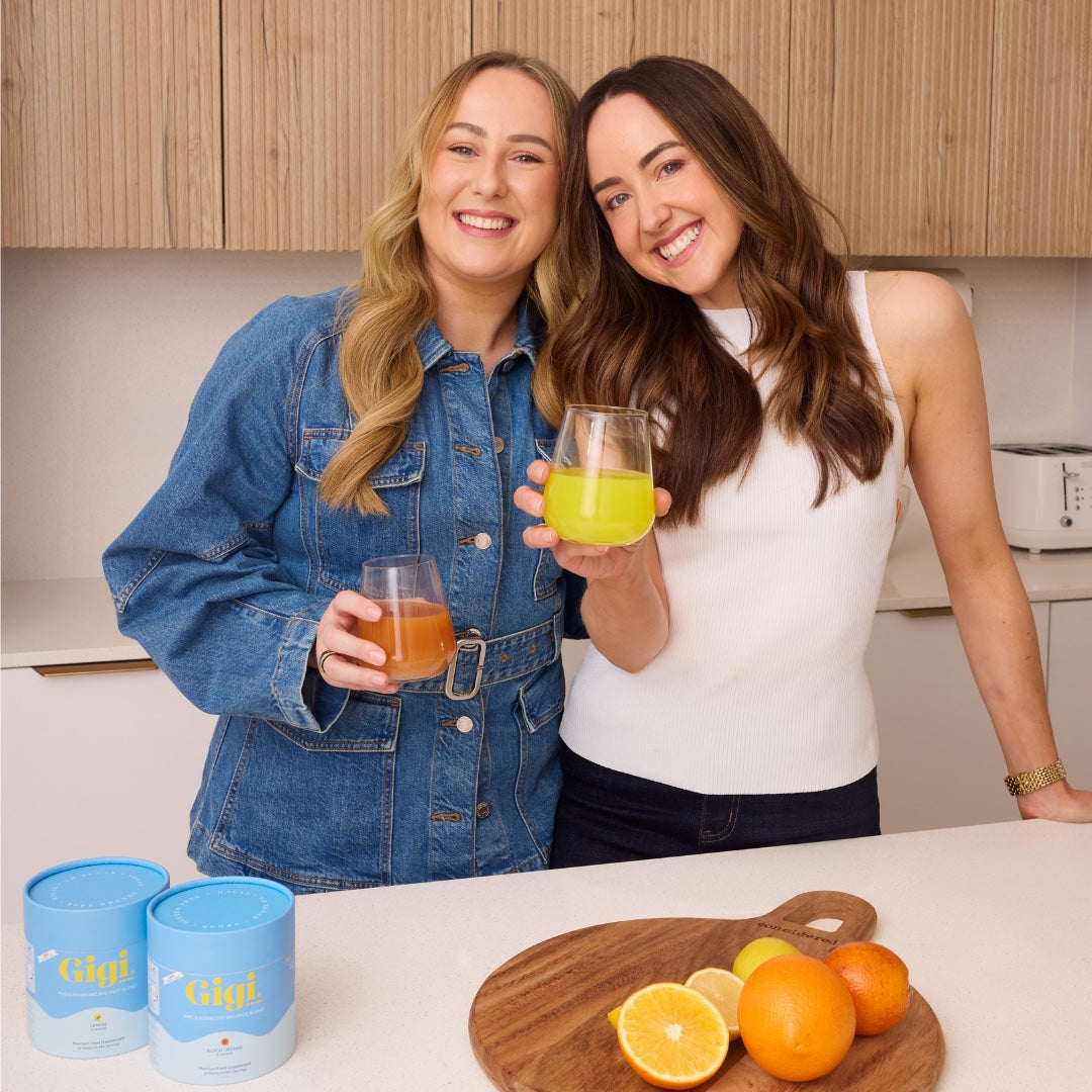 Two smiling women in a kitchen holding Gigi-mixed drinks beside both PMS & Hormone Balance Blend flavours